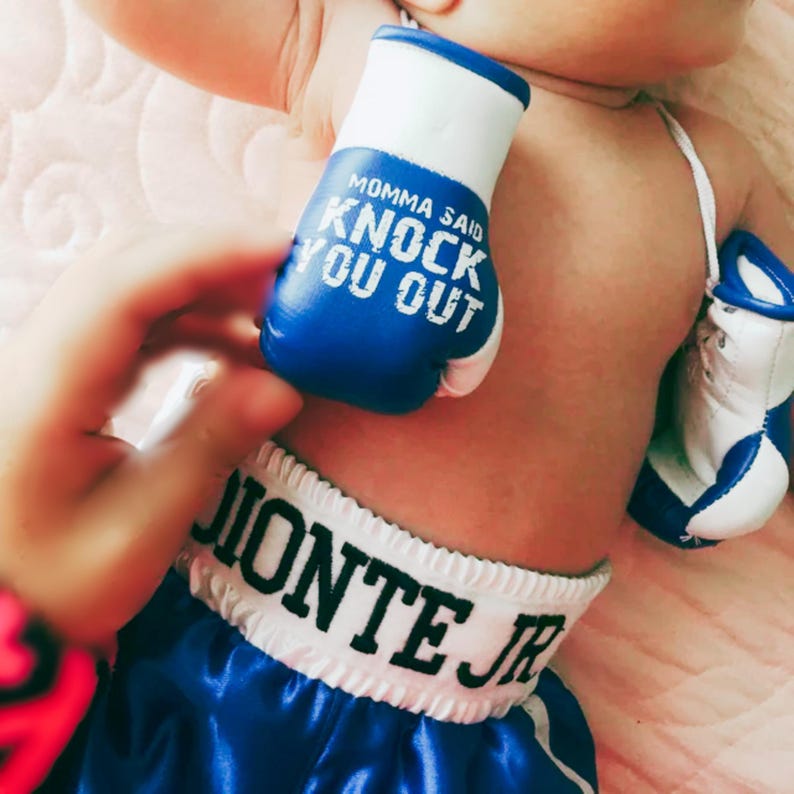 Pu&ograve; includere: Un bambino indossa pantaloncini da boxe blu satinati con il nome "JIONTE JR" sulla cintura. Il bambino indossa anche guantoni da boxe blu e bianchi con la scritta "MOMMA SAID KNOCK YOU OUT".