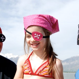 May include: Three pirate-themed head coverings and eye patches. One red bandana with a black eye patch, one pink polka-dot bandana with a pink skull eye patch, and one black bandana with a white skull and crossbones print and a black eye patch.