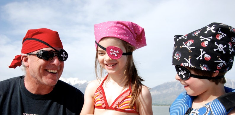 May include: Three pirate-themed head coverings and eye patches. One red bandana with a black eye patch, one pink polka-dot bandana with a pink skull eye patch, and one black bandana with a white skull and crossbones print and a black eye patch.