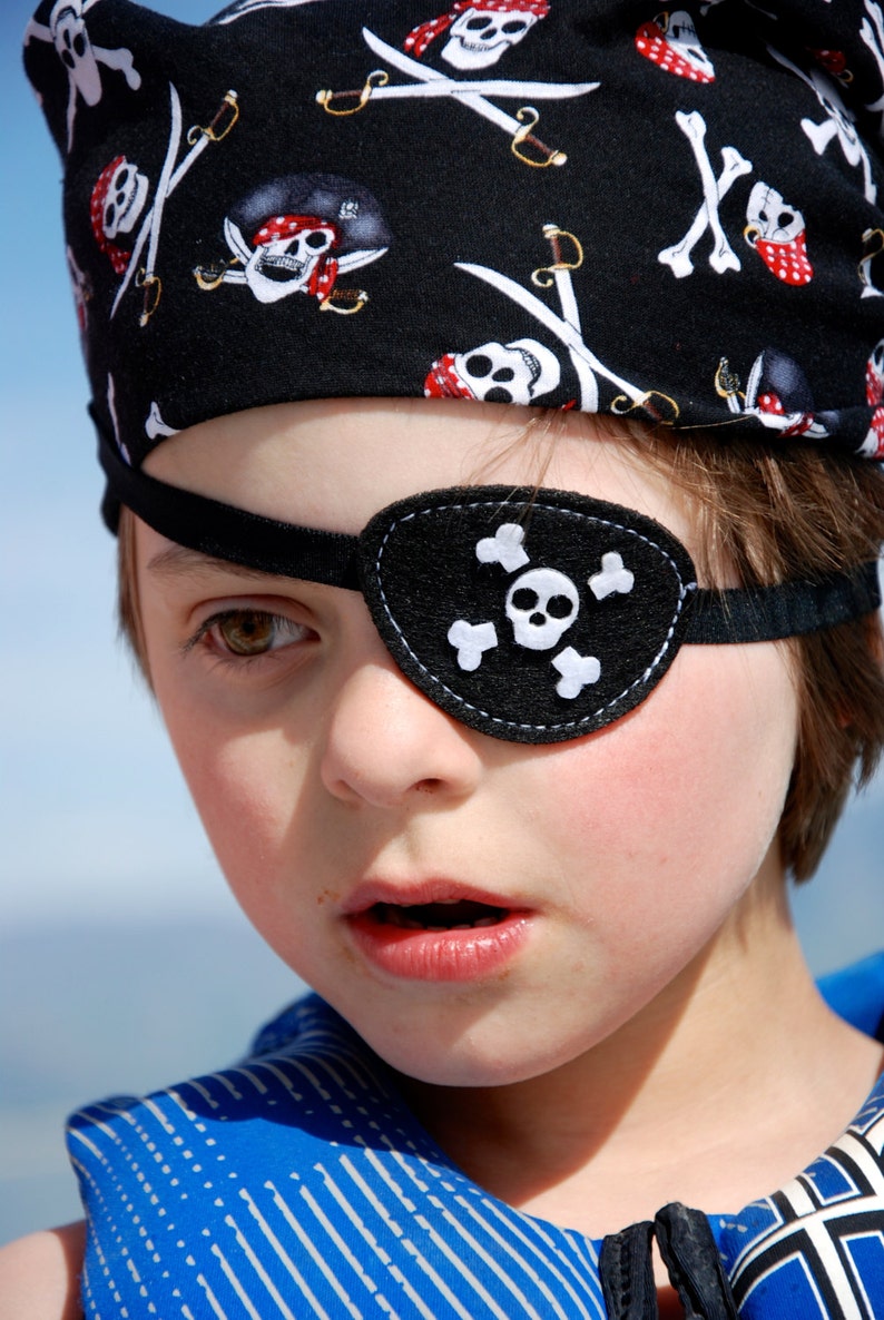 May include: A black pirate-themed eye patch featuring a white skull and crossbones motif, secured with a black elastic strap. The eye patch is worn over one eye, complemented by a matching black bandana patterned with white skulls and swords.