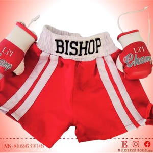 K&ouml;nnte beinhalten: Rote und wei&szlig;e Boxershorts mit dem Namen "BISHOP" auf dem Bund gestickt. Die Shorts werden mit einem Paar wei&szlig;en Boxhandschuhen geliefert, auf denen der Text "Lil Champ" gestickt ist.