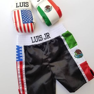 May include: Black satin boxing shorts with American and Mexican flag accents. The shorts have the name "LUIS JR" embroidered on the waistband. Two white boxing gloves with the names "LUIS" and "JR" and the American and Mexican flags are also pictured.