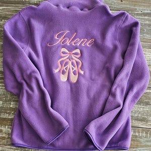 May include: A purple fleece pullover jacket featuring long sleeves and a high collar. The back of the garment is embroidered with the name Jolene in pink script lettering above a detailed pink graphic of ballet slippers tied with a bow.