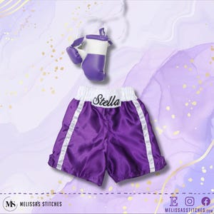K&ouml;nnte beinhalten: Lilafarbene Boxershorts aus Satin mit wei&szlig;em Besatz und dem Namen "Stella" auf der Taille gestickt. Die Shorts werden mit einem Paar lila und wei&szlig;en Boxhandschuhen geliefert.