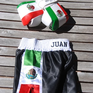 May include: Black and green boxing shorts with a white waistband and the word "JUAN" embroidered on the waistband. The shorts have a Mexican flag design with a green, white, and red color scheme.