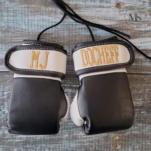 May include: Pair of miniature boxing gloves in black and white with gold embroidered text. One glove reads "MJ" and the other reads "DOCHEFF". Each glove has a white wrist strap and a black cord for hanging.