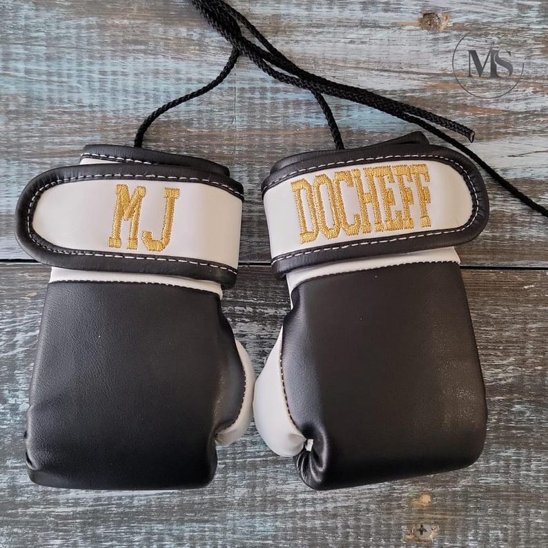 May include: Pair of miniature boxing gloves in black and white with gold embroidered text. One glove reads "MJ" and the other reads "DOCHEFF". Each glove has a white wrist strap and a black cord for hanging.