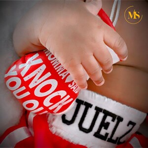 Pu&ograve; includere: Un guantone da boxe rosso e bianco con la scritta "KNOCK YOU OUT" e "MOMMA SAID" in lettere bianche. Il guantone &egrave; tenuto dalla mano di un bambino. Sono visibili anche pantaloncini da boxe con il nome "JUELZ".
