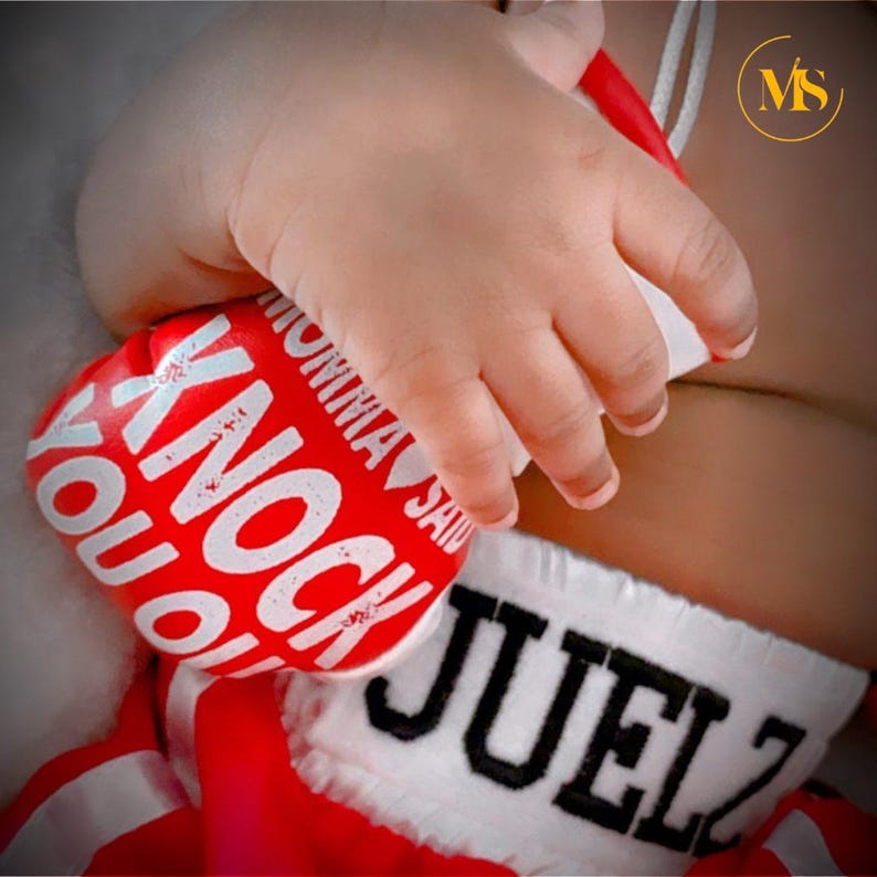 Pu&ograve; includere: Un guantone da boxe rosso e bianco con la scritta "KNOCK YOU OUT" e "MOMMA SAID" in lettere bianche. Il guantone &egrave; tenuto dalla mano di un bambino. Sono visibili anche pantaloncini da boxe con il nome "JUELZ".