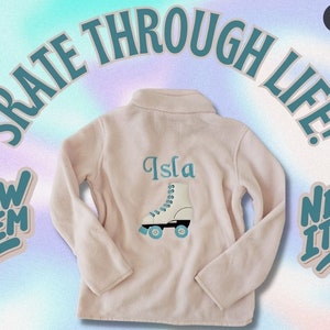 Puede incluir: Chaqueta de forro polar rosa claro con cuello con cremallera. La parte trasera presenta el nombre "Isla" sobre un patín blanco con detalles azules. Las palabras "Skate Through Life!" están arqueadas arriba, con pegatinas "New Item" a cada lado.