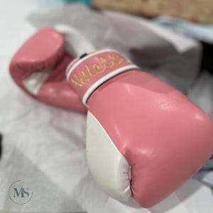 May include: Pink boxing gloves with white accents. The gloves have the name "Natalie" embroidered in gold thread on the wrist strap. The gloves are made of a smooth material and are designed for boxing or other combat sports.