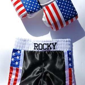 May include: Black boxing shorts with red, white, and blue American flag stripes on the sides. The waistband is white with the word "ROCKY" embroidered in black. The shorts are paired with a set of white boxing gloves with red, white, and blue American flag designs and the word "USA" printed on the gloves.