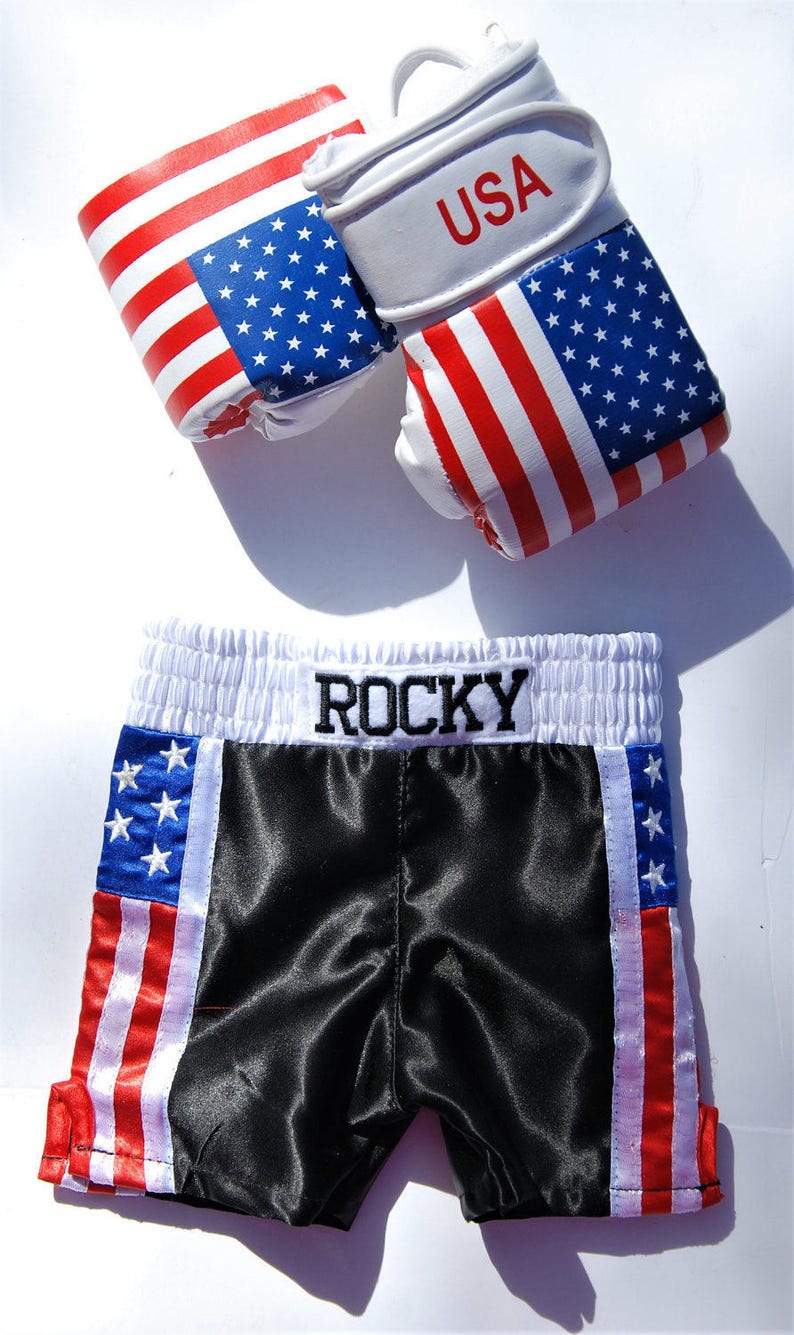 May include: Black boxing shorts with red, white, and blue American flag stripes on the sides. The waistband is white with the word "ROCKY" embroidered in black. The shorts are paired with a set of white boxing gloves with red, white, and blue American flag designs and the word "USA" printed on the gloves.