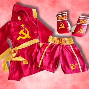 May include: A red boxing robe, shorts, and gloves set. The robe has a hood, yellow trim, and a yellow hammer and sickle symbol. The shorts and gloves also feature the symbol and the name "DRAGO".