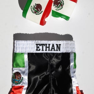 May include: Black satin boxing shorts with green, white, and red trim. The shorts have the Mexican flag on the side and the name "ETHAN" embroidered on the waistband.