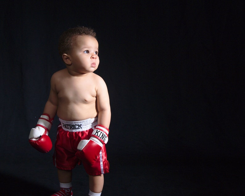 May include: A young boy wearing red boxing shorts with white trim and the words "Patrick King" on the waistband. He is wearing red and white boxing gloves and looking to the right.
