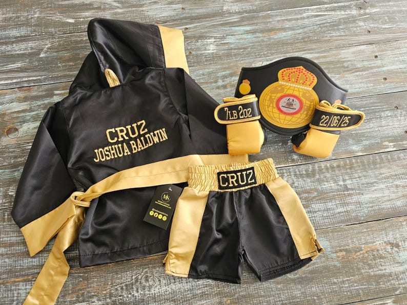 May include: Black and gold boxing outfit set. Includes a hooded robe with "CRUZ JOSHUA BALDWIN" in gold lettering, boxing shorts, gloves with "7LB 2OZ" and "22/06/25" and a championship belt.