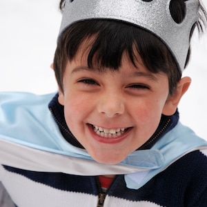 May include: A young boy wearing a silver crown and a blue and white striped sweater with a blue cape. He is smiling and looking at the camera.