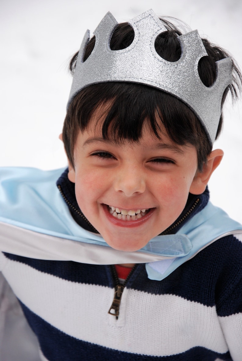 May include: A young boy wearing a silver crown and a blue and white striped sweater with a blue cape. He is smiling and looking at the camera.