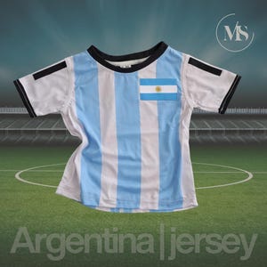 May include: A white and light blue striped Argentina jersey with a black collar and sleeve trim. The jersey features the Argentina flag on the chest and the text "Argentina Jersey" at the bottom.