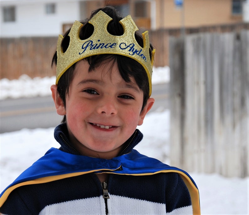 May include: A young boy wearing a gold crown with the text "Prince Ayden" on it. He is wearing a blue and white striped shirt with a blue cape.