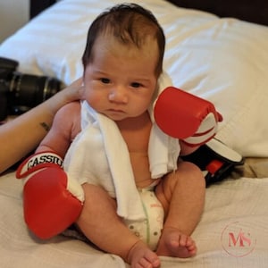 Peut inclure: Un bébé portant des gants de boxe rouges et blancs, avec le nom "Cassius" sur l'un d'eux. Le bébé porte également une serviette blanche autour du cou et une couche. Le bébé est assis sur un lit blanc.