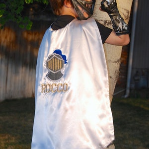 May include: A child's knight costume featuring a silver helmet, black gauntlets, and a white satin cape. The cape is embroidered with a shield design and the name "ROCCO" in gold thread. The child is wearing camouflage pants.