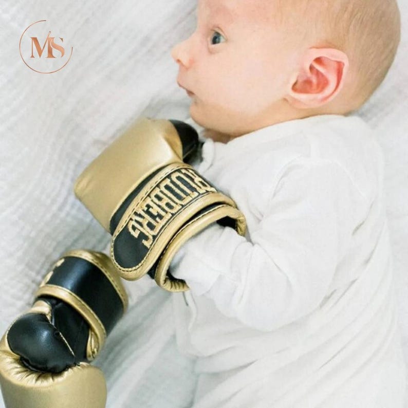 May include: A pair of small gold and black boxing gloves with the word "RODBERG" in gold lettering. The gloves are resting on a white surface.