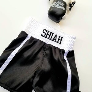 K&ouml;nnte beinhalten: Schwarze Satin-Boxershorts mit wei&szlig;em Besatz und dem Namen "SHIAH" auf dem Bund gestickt. Ein Paar schwarz-wei&szlig;e Boxhandschuhe mit dem Text "FUTURE CHAMP" sind ebenfalls abgebildet.