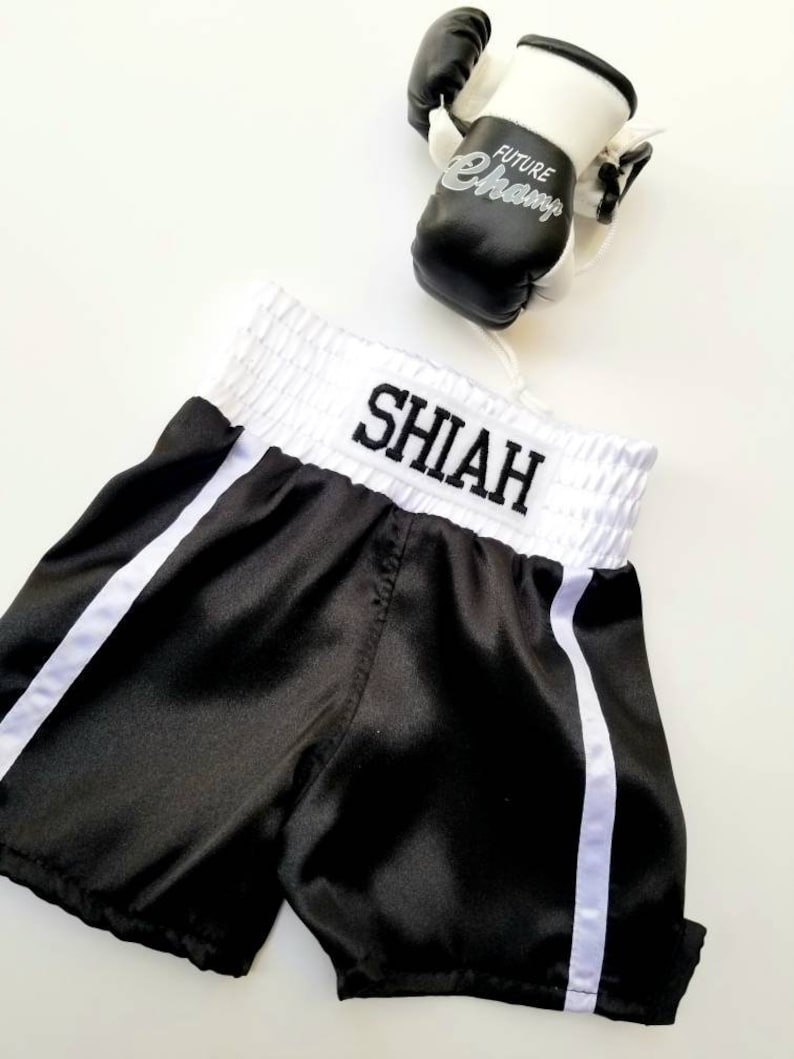 K&ouml;nnte beinhalten: Schwarze Satin-Boxershorts mit wei&szlig;em Besatz und dem Namen "SHIAH" auf dem Bund gestickt. Ein Paar schwarz-wei&szlig;e Boxhandschuhe mit dem Text "FUTURE CHAMP" sind ebenfalls abgebildet.