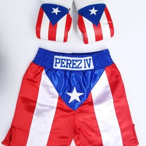 May include: A red, white, and blue boxing shorts set with the text "PEREZ IV" embroidered on the waistband. The shorts have a blue satin panel with a white star in the center. The set also includes a pair of white boxing gloves with the Puerto Rican flag printed on them.