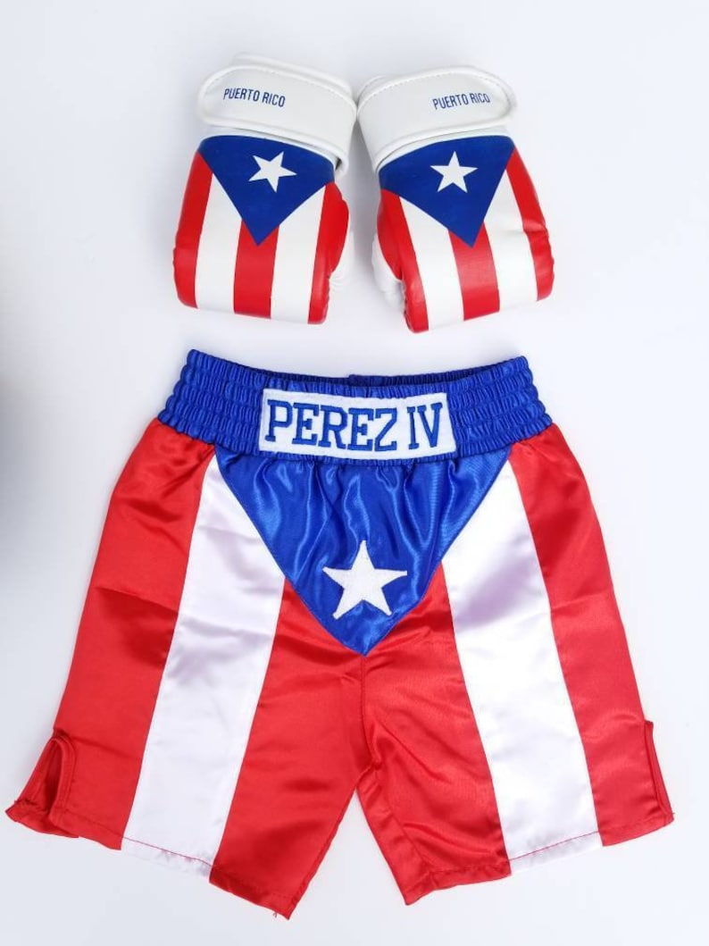May include: A red, white, and blue boxing shorts set with the text "PEREZ IV" embroidered on the waistband. The shorts have a blue satin panel with a white star in the center. The set also includes a pair of white boxing gloves with the Puerto Rican flag printed on them.