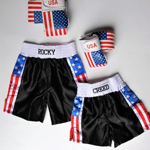 May include: Two pairs of black satin boxing shorts with red, white, and blue American flag accents. One pair says "ROCKY" and the other says "CREED".