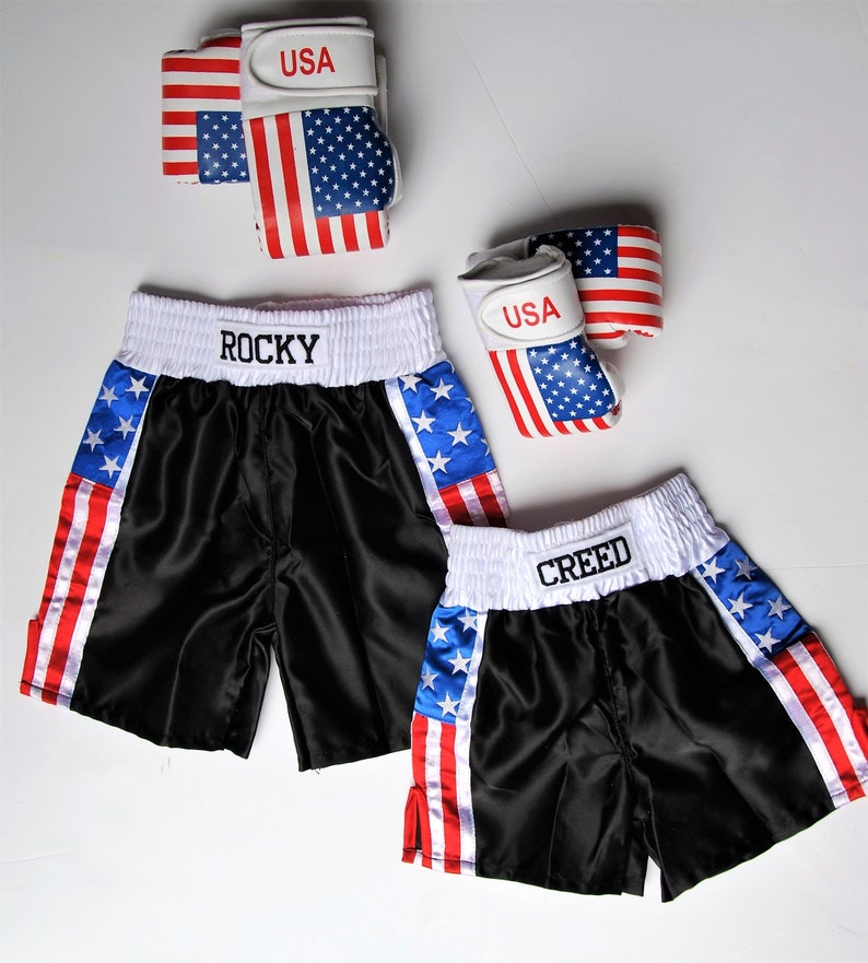 May include: Two pairs of black satin boxing shorts with red, white, and blue American flag accents. One pair says "ROCKY" and the other says "CREED".
