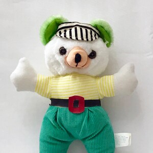 May include: White plush teddy bear wearing a striped hat with green ears, a yellow and white striped shirt, a black belt with a red buckle, and green pants.
