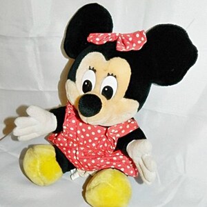 K&ouml;nnte beinhalten: Ein Pl&uuml;schtier von Minnie Mouse, das ein rotes Kleid mit wei&szlig;en Punkten und eine rote Schleife mit wei&szlig;en Punkten tr&auml;gt. Das Spielzeug hat schwarze Ohren, eine schwarze Nase und gelbe Schuhe.