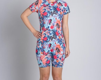 Könnte beinhalten: Ein einteiliger Badeanzug mit kurzen Ärmeln und einem lebendigen Blumen- und Schmetterlingsmuster. Der Badeanzug hat einen hellblauen Hintergrund mit rosa, roten und lila Blumen und Schmetterlingen. Das Kleidungsstück hat einen hohen Ausschnitt und kurze Shorts.