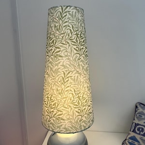 May include: A tall table lamp with a cylindrical lampshade. The shade features a repeating pattern of green and beige leaves. The base of the lamp is a rounded, light blue color. The lamp is turned on, emitting a soft glow.