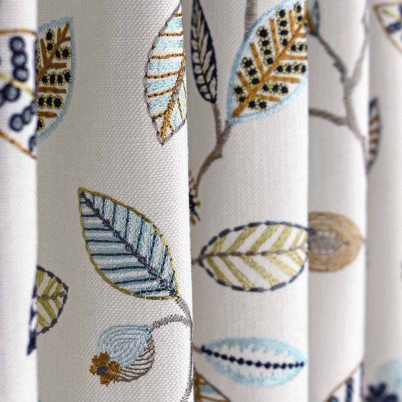 May include: White fabric with an embroidered pattern of blue, green, and gold leaves. The leaves are arranged in a repeating pattern.