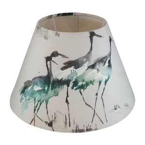 May include: A conical lampshade featuring a watercolour illustration of three cranes in teal, green, and black. The birds have long legs and necks, set against a neutral backdrop. The lampshade has a linen-like texture, suitable for a table lamp.
