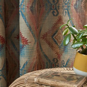 May include: A close-up of a woven curtain with a geometric pattern in shades of blue, red, and brown. A small potted plant with green leaves sits on a woven table with a book on top.