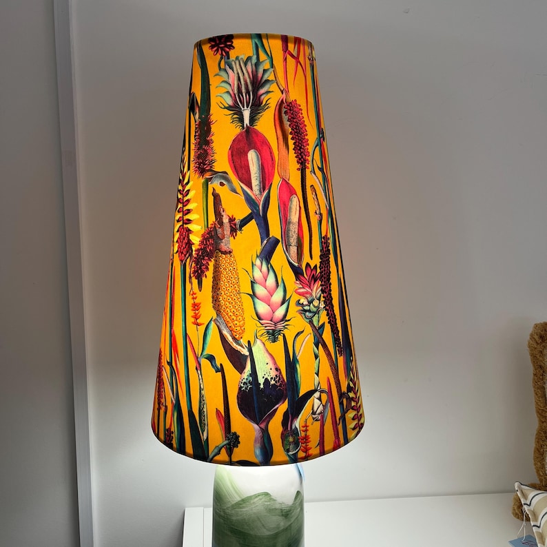 May include: A tall lampshade with a vibrant yellow background and a botanical print featuring various colorful flowers and plants. The lampshade is cone-shaped and sits atop a green and white glass base, creating a decorative lighting fixture.