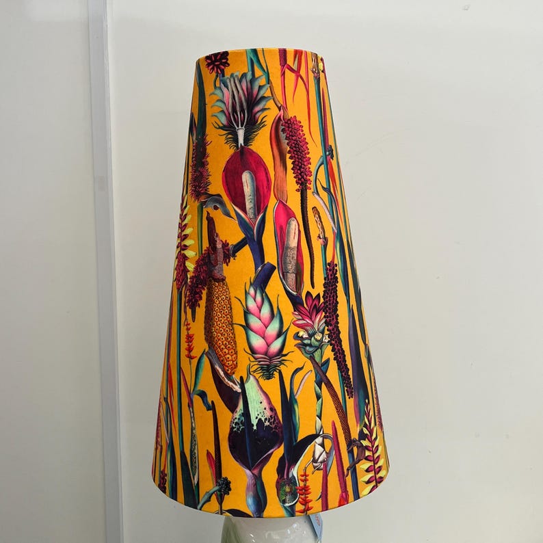 May include: A tall, conical lampshade with a vibrant botanical print. The shade features a mustard yellow background with colorful illustrations of various plants and flowers in shades of red, green, purple, and pink. The lampshade is a decorative lighting element.