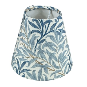 May include: A tapered drum lampshade featuring a white background with a repeating botanical pattern of willow leaves in various shades of blue and muted brown. The fabric has a visible woven texture, and the interior is lined with white material.