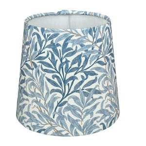 May include: A cylindrical lampshade with a botanical pattern. The design features blue and grey leaves and branches against a white background. The lampshade is a light fixture for a table lamp or floor lamp.