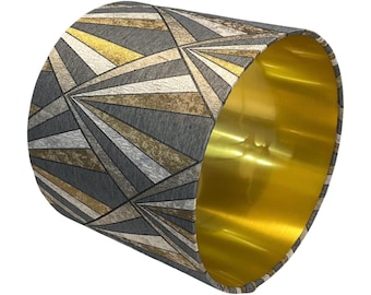 May include: A cylindrical lampshade with a geometric pattern in shades of gray, gold, and cream. The interior of the lampshade is a bright gold color. The design features a repeating pattern of angled lines and shapes.
