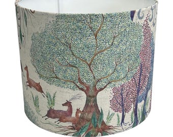 May include: Cylindrical lampshade featuring a forest scene with a large tree, deer, and other trees in shades of green, brown, and purple. The interior is white. The lampshade is designed to fit over a lamp base.