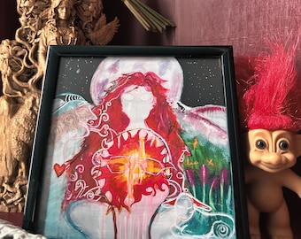 May include: A framed painting featuring a central figure with long red hair, a glowing orange sun-like sphere at the chest, and a large moon in the background. The artwork includes white line art of a cat and swirling patterns against a dark sky.
