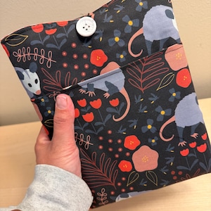 May include: A black fabric book sleeve with a floral and opossum pattern. The design features red, pink, and yellow flowers, with opossums in gray and pink. A white button secures the flap. The sleeve is held in a person's hand.