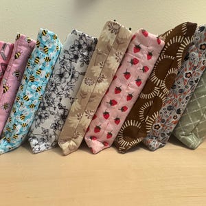 May include: Nine fabric pouches with different patterns, including bees, flowers, strawberries, and geometric designs. The pouches are lined up in a row on a light wood surface.
